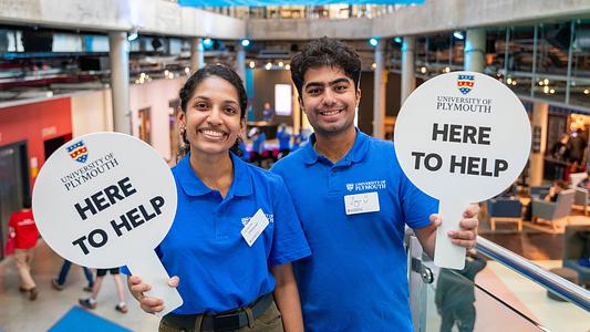Two student ambassadors holding 'here to help' signs at an undergraduate open day.